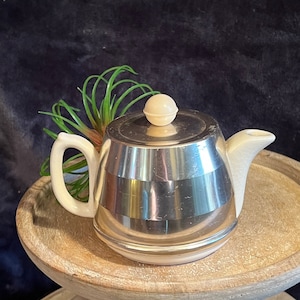 May include: A silver teapot with a white lid and a white handle. The teapot is sitting on a wooden pedestal.