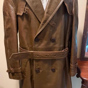 May include: A double-breasted brown leather trench coat featuring a wide collar, shoulder epaulets, a matching waist belt with a buckle, and adjustable straps on the cuffs. The coat has four dark brown buttons and a smooth, polished finish.