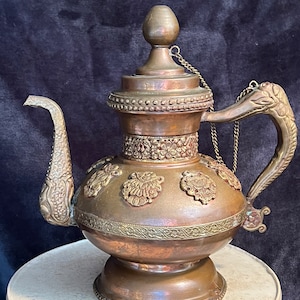 May include: An antique copper teapot with ornate detailing. The teapot features a curved spout, a decorative handle, and a lid with a small knob. The body is adorned with embossed floral patterns and a decorative band around the middle. A chain connects the lid to the handle.