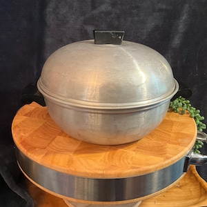 May include: A round, brushed aluminum cooking pot with a matching domed lid. The pot features two black side handles and a black knob on the center of the lid. The metal surface shows a matte finish with subtle signs of wear and light scratches.
