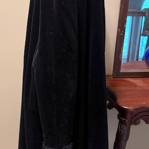May include: A long, black velvet coat featuring a high collar and long sleeves with textured, dark gray fuzzy cuffs. The coat has a smooth, matte black finish and a straight, floor-length silhouette that hangs vertically from the shoulders.
