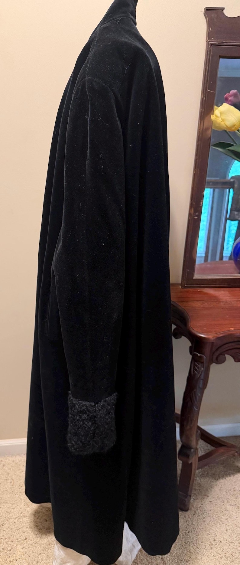 May include: A long, black velvet coat featuring a high collar and long sleeves with textured, dark gray fuzzy cuffs. The coat has a smooth, matte black finish and a straight, floor-length silhouette that hangs vertically from the shoulders.