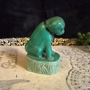 May include: A vintage, jade green ceramic dog figurine. The dog is sitting on a tree stump base and wearing a black collar. The figurine has a glossy finish and is a decorative item.