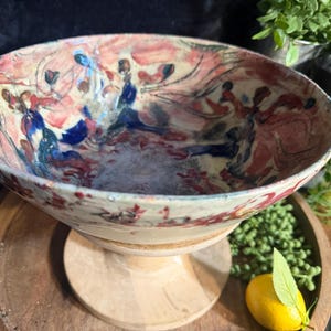 Może przedstawiać: Ceramiczna miska z szerokim, rozszerzającym się brzegiem, ozdobiona abstrakcyjnym, malarskim wzorem w odcieniach różu, błękitu i ciemnej czerwieni na kremowym tle. Misa osadzona jest na jasnej, gładkiej drewnianej podstawie z okrągłą stopką.