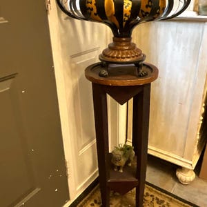May include: A decorative black metal and bronze pedestal with a round top and a decorative bowl. The bowl has a black cage design with yellow objects inside. The pedestal has a dark brown finish and a small shelf with a figurine.