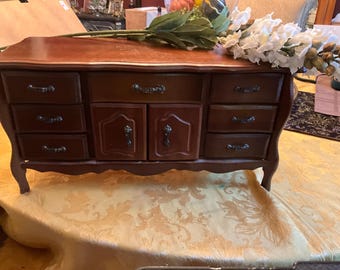 Puede incluir: Un joyero de madera con acabado marrón oscuro. Cuenta con múltiples cajones y puertas de armario con tiradores ornamentados. La caja descansa sobre un mantel estampado, con un arreglo floral encima.