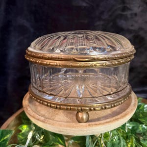 May include: A round glass trinket box featuring a clear, faceted, and ribbed design. The container is encased in a decorative antique brass-toned metal frame with a matching lid rim and three small, spherical brass-toned feet at the base.