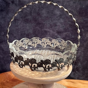 May include: A clear glass bowl with a scalloped edge and a silver metal handle. The bowl is decorated with a floral design and sits on a wooden pedestal.