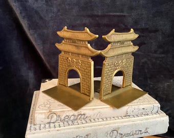 May include: A pair of gold-toned metal bookends shaped like traditional Asian temple gates. The bookends are detailed with intricate carvings and have a tiered roof design.