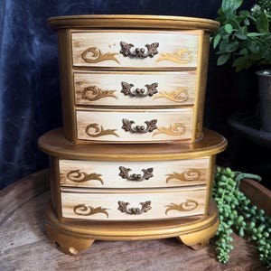 May include: A two-tiered jewelry chest with a gold frame and five drawers. Each drawer features a cream-colored front with gold-painted scrollwork accents and small, ornate, dark metal handles. The chest has a curved, oval-shaped design.