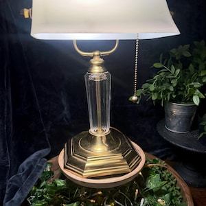 May include: A banker's lamp featuring a rectangular white glass shade, a clear fluted glass column, and a polished brass base. The lamp includes a brass pull chain for operation and emits a bright light from beneath the shade.