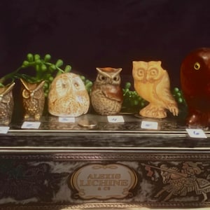 May include: A collection of six owl figurines, each with unique details and colors. The owls are arranged in a row on a wooden surface with a label that reads "Alexis Lichine & Cie".