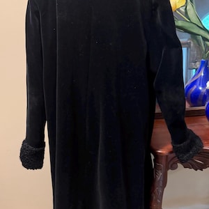 May include: A long, black coat featuring a high collar and long sleeves with textured, black faux-fur cuffs. The coat is displayed on a mannequin, showing a smooth, solid black fabric construction that extends to a mid-calf length.