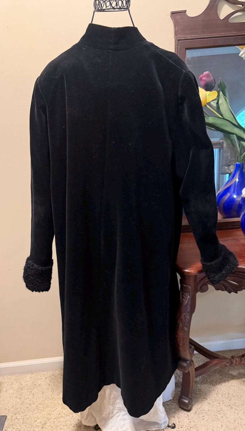 May include: A long, black coat featuring a high collar and long sleeves with textured, black faux-fur cuffs. The coat is displayed on a mannequin, showing a smooth, solid black fabric construction that extends to a mid-calf length.