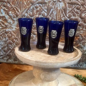 May include: Four cobalt blue shot glasses with silver metal accents. Each glass has the number "247" printed on the front. The glasses are arranged on a wooden pedestal.