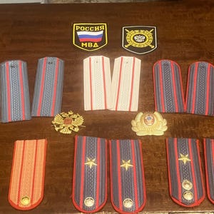 May include: A collection of military insignia patches and badges, including a Russian flag patch with the text "Россия МВД", a black patch with a gold emblem, and several sets of shoulder boards with red and blue stripes.