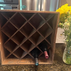 May include: A square wooden wine rack with a diamond-shaped grid pattern for bottle storage. The rack features a natural wood finish and includes two wine bottles placed in the bottom slots. The text Great Western 1860 is printed on the base.