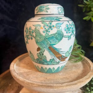 May include: A white ceramic ginger jar with a lid, featuring a hand-painted design of two teal birds perched on branches with green floral accents. The surface has a visible crackle glaze finish and a base decorated with green leaf motifs.