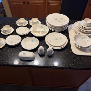 May include: A vintage mid-century modern dinnerware set. The set includes plates, bowls, cups, saucers, and serving dishes. The white ceramic pieces feature a blue and yellow starburst design. Salt and pepper shakers and a butter dish are also included.