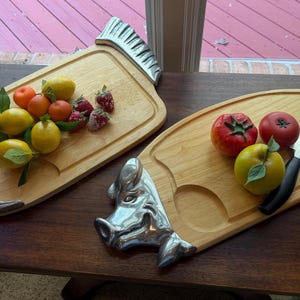 May include: Two light-colored wooden serving boards with metallic handles. One board features a fish-shaped handle and holds lemons, oranges, and strawberries. The other board has a pig-shaped handle and holds two tomatoes, a yellow apple, and a knife.