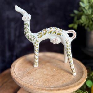 May include: A white ceramic llama figurine featuring hand-painted green and brown leaf patterns along its neck, back, and legs. The stylized animal has a long neck, a small tail, and a delicate floral design painted on its side.