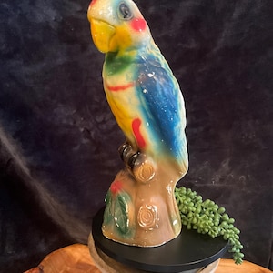 May include: A colorful ceramic parrot figurine perched on a tree stump. The parrot features a yellow beak, red accents, and blue, yellow, and green plumage. The figurine is mounted on a black base.