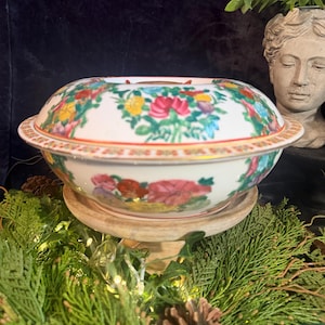 Puede incluir: Un cuenco de servicio redondo de porcelana con tapa, que presenta una base blanca decorada con vibrantes motivos florales en tonos rojo, amarillo, verde y azul, acentuado por un borde dorado fino y un patrón geométrico en la tapa.