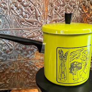 May include: A yellow enamel saucepan with a black handle and lid. The saucepan has a black and white graphic of asparagus, green beans, and broccoli on the side. The text "ASPARAGUS" and "BROCCOLI" are visible on the graphic.