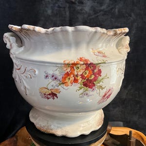 May include: A large, antique white ceramic planter with a scalloped rim and floral design. The planter features a pedestal base and handles. The floral design includes red, orange, and purple flowers. The rim and base have a gold-colored trim.