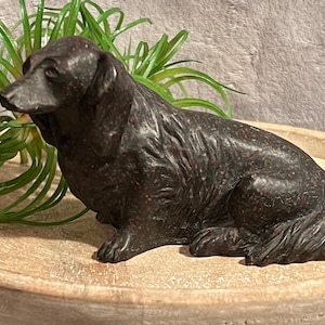 May include: A black resin sculpture of a dog sitting with its head turned to the side. The sculpture is detailed with textured fur and is sitting on a wooden surface.