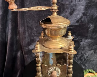 May include: An antique brass coffee grinder with a wooden handle and knob. The grinder has a decorative floral design on the front and is set on a black surface. The grinder is a vintage kitchenware item.