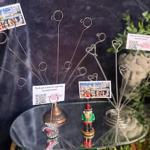 May include: Two metal wire card holders with multiple stems featuring circular and heart-shaped clips. One holder has a bronze base, the other a silver spiral base. A small nutcracker figurine stands at the base of one, holding a business card.