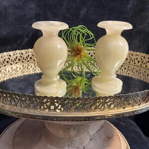 Puede incluir: Dos jarrones de ónix color crema con forma clásica sobre una bandeja espejada con borde decorativo dorado. Una pequeña planta con hojas verdes y marrones se encuentra entre los jarrones. La bandeja está sobre un pedestal marmóreo.