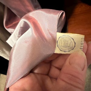 May include: A light pink satin lining inside a garment with a cream-colored fabric tag. The tag features a circular blue logo with the text National Coat and Suit Industry Recovery Board and the number 81677 printed on the side.