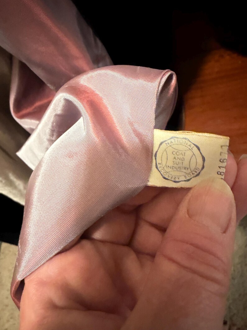 May include: A light pink satin lining inside a garment with a cream-colored fabric tag. The tag features a circular blue logo with the text National Coat and Suit Industry Recovery Board and the number 81677 printed on the side.