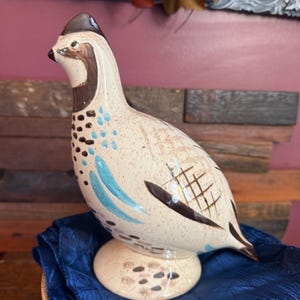 Könnte beinhalten: Keramik-Wachtelfigur mit beigem Körper, braunen und schwarzen Markierungen und blauen Akzenten. Der Vogel hat einen dunkelbraunen Kamm und sitzt auf einem runden Sockel. Die Figur wird auf einem blauen Tuch präsentiert.