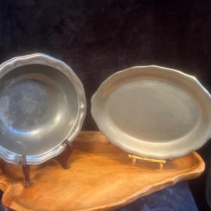May include: Two silver-colored pewter serving dishes, one round and one oval, are displayed on a wooden surface. Both dishes have a slightly raised rim.