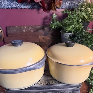 May include: Two vintage yellow enamel pots with lids. The pots have a silver trim and black handles. The pots are sitting on a decorative box. The background includes plants and a vintage clock.