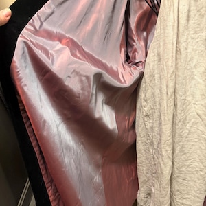 May include: A black coat with a smooth, iridescent mauve-pink lining. The coat is open, revealing the interior fabric, and is positioned next to a textured, light beige garment with a ruffled hemline visible on the right side.