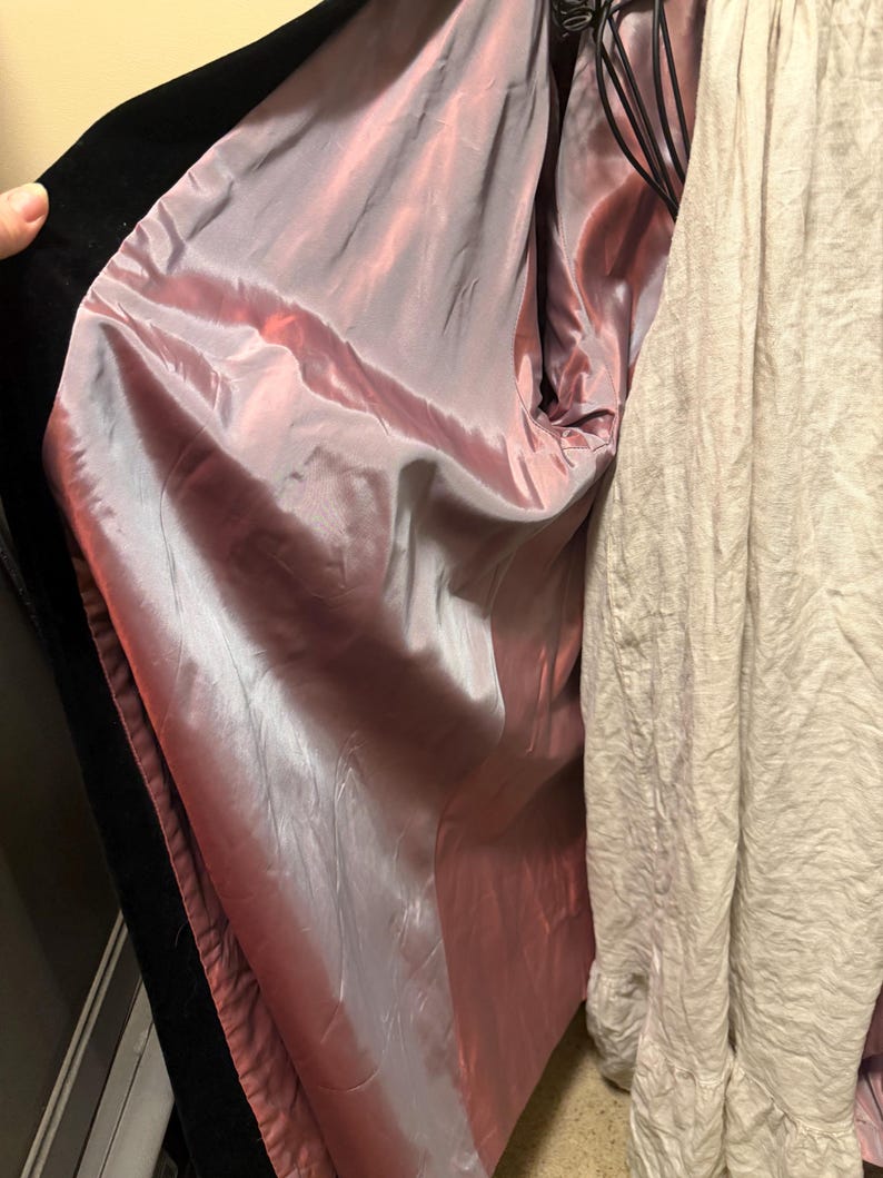 May include: A black coat with a smooth, iridescent mauve-pink lining. The coat is open, revealing the interior fabric, and is positioned next to a textured, light beige garment with a ruffled hemline visible on the right side.