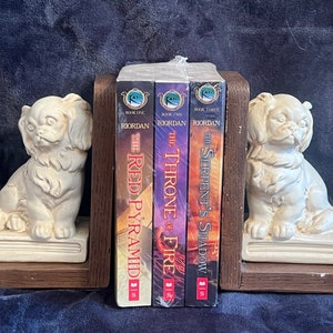 May include: A set of bookends featuring two white ceramic dog figurines. The bookends are holding up three paperback books with the titles "The Red Pyramid", "The Throne of Fire", and "The Serpent's Shadow".