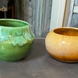 May include: Two ceramic bowls. One is a vibrant green with a glossy finish, the other is a warm yellow-brown. Both have a rounded shape with a wide opening, suitable for decorative or functional use. They sit on a dark surface.