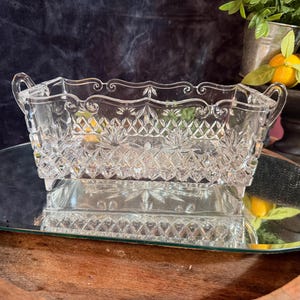 Può includere: Un piatto rettangolare in vetro trasparente con un bordo smerlato decorativo, due manici laterali e un motivo a rombi pressato sui lati. La base del piatto presenta un design a stella e il vetro ha una consistenza sfaccettata.
