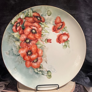 May include: A large, white porcelain plate with a gold rim features a hand-painted design of red poppies with green leaves and stems. The plate is on a stand.