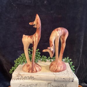 May include: Two ceramic deer figurines with a brown and white marbled glaze. The deer are standing on a stack of books.
