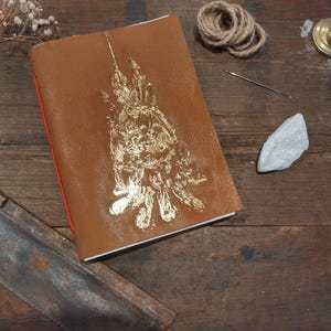 May include: A brown leather journal with gold foil embellishments. The journal is open, revealing red-edged pages. A coil of twine, a needle, and a white stone are arranged on a wooden surface, along with a small saw.