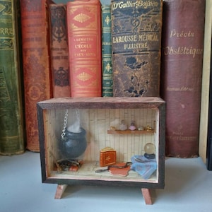 Puede incluir: Un televisor de madera en miniatura con un diorama en su interior. El diorama presenta un caldero negro con vapor saliendo de él, un libro, una bola de cristal y otros objetos mágicos.