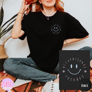 May include: Black t-shirt with a circular design featuring a smiling face and the words "OCCUPATIONAL THERAPY". The design is in light blue and is also on a black square. The shirt is a casual style.