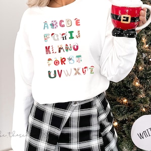 May include: White sweatshirt featuring a Christmas alphabet design. Each letter is adorned with festive decorations such as candy canes, Santa hats, and baubles. The sweatshirt is worn with black and white checked trousers.