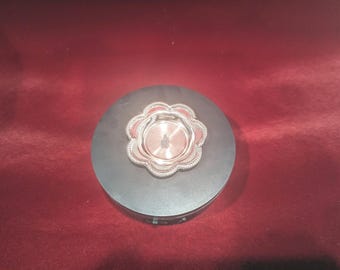 May include: A silver-colored, decorative dish with a scalloped edge sits on a black circular base. The dish has a raised center and a smooth, reflective surface.
